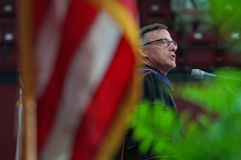 President Kevin O'Brien speaks at his inauguration. His face is framed by a flag and plant.