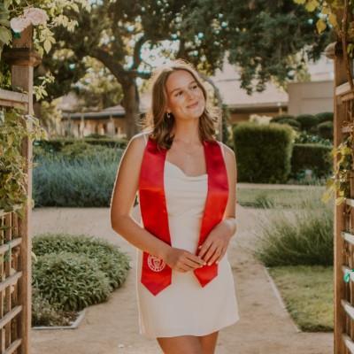 A person posing outdoors wearing a white dress and red stole.