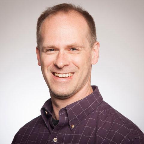 A person smiling, wearing a dark button-up shirt against a light background.