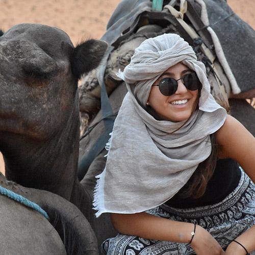 A person sitting beside a camel, wearing sunglasses and headscarf.