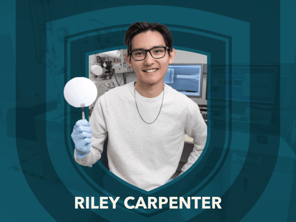 A male student wearing medical-grade gloves holds a silicon disk in front of a large microscope and screens filled with magnified images in a lab setting.