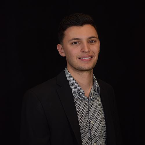A person in a dark suit with a patterned shirt smiles against a black background.
