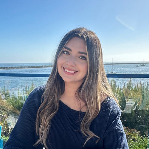 Alexis sits at an outdoor dining table with the water and sky in the background