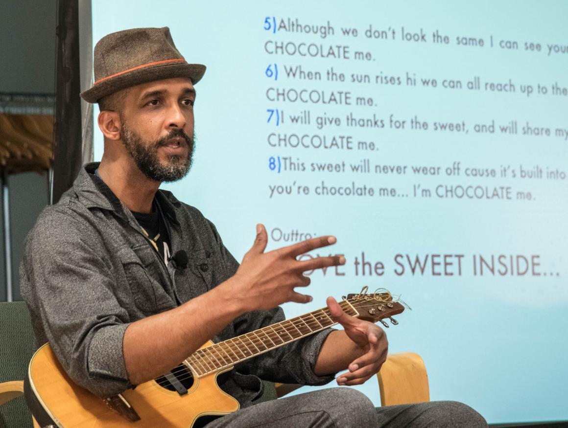 Shane Evans in front of screen with guitar in lap gesturing with hands 