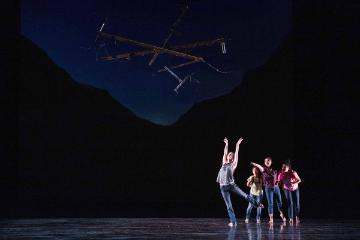Dancers performing on stage with a dark background and minimalist lighting.