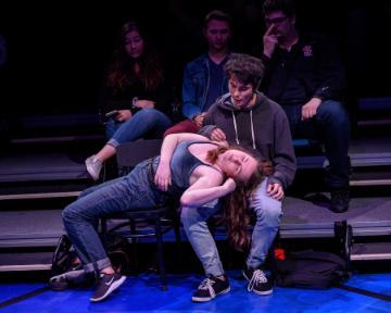 Two actors perform an emotional scene on stage, with a few onlookers behind them.