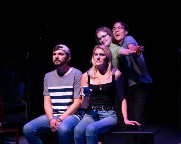 Four people on a dark stage; two seated, two standing.