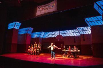 Alt text: Contestant on stage in a spelling bee competition with judges and audience.