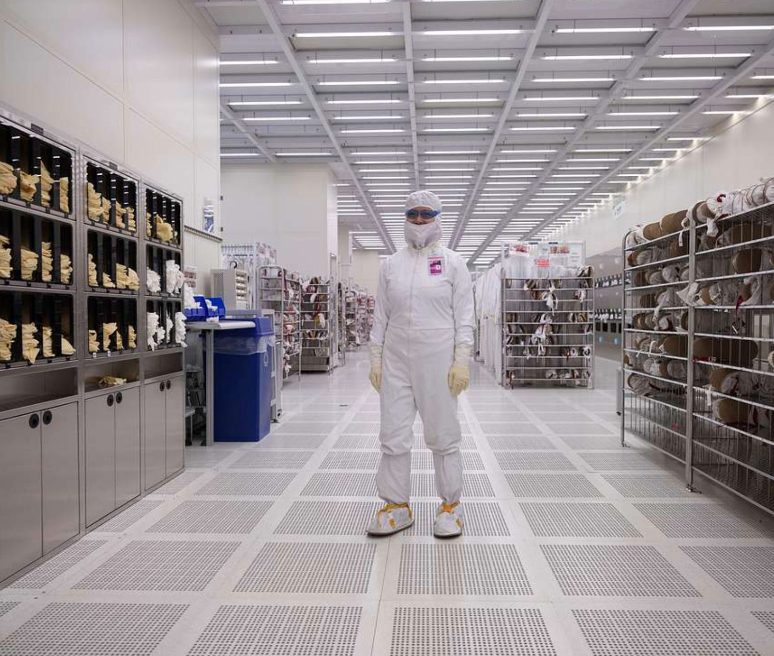Person in protective clothing stands in a cleanroom with shelves and equipment.