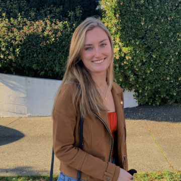 A woman standing outdoors, smiling, wearing a brown jacket.