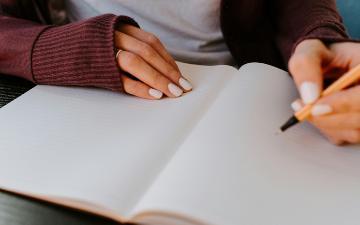 Woman's hands at a desk writing in a blank notebook. Photo by Photo by Kelly Sikkema on Unsplash.