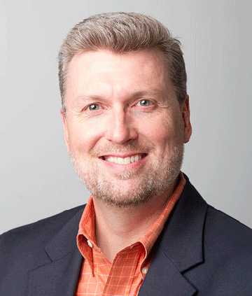 A man smiling, wearing an orange shirt and dark blazer.