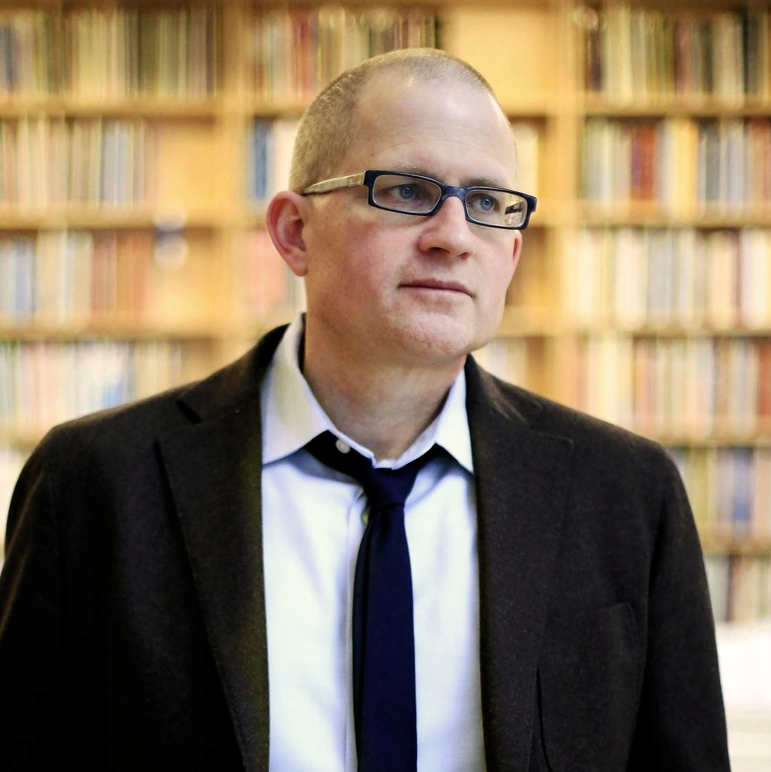 A person in a suit stands in front of bookshelves.