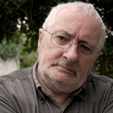 Man with glasses tilting his head outdoors next to dark green leaves.