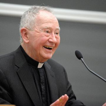 A person in clerical attire speaking at a microphone, smiling.