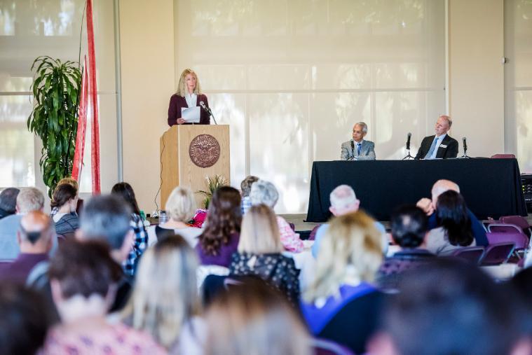People attending a climate conference, one speaker at a podium, two panelists seated.