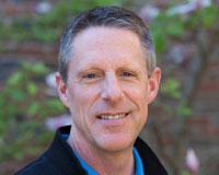 A person smiling outdoors with a blurred natural background.