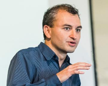 A person speaking and gesturing in front of a blank screen.