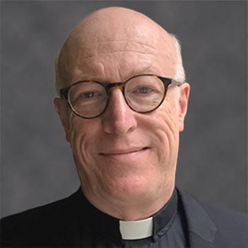 A balding man in clerical attire and glasses smiles at the camera.