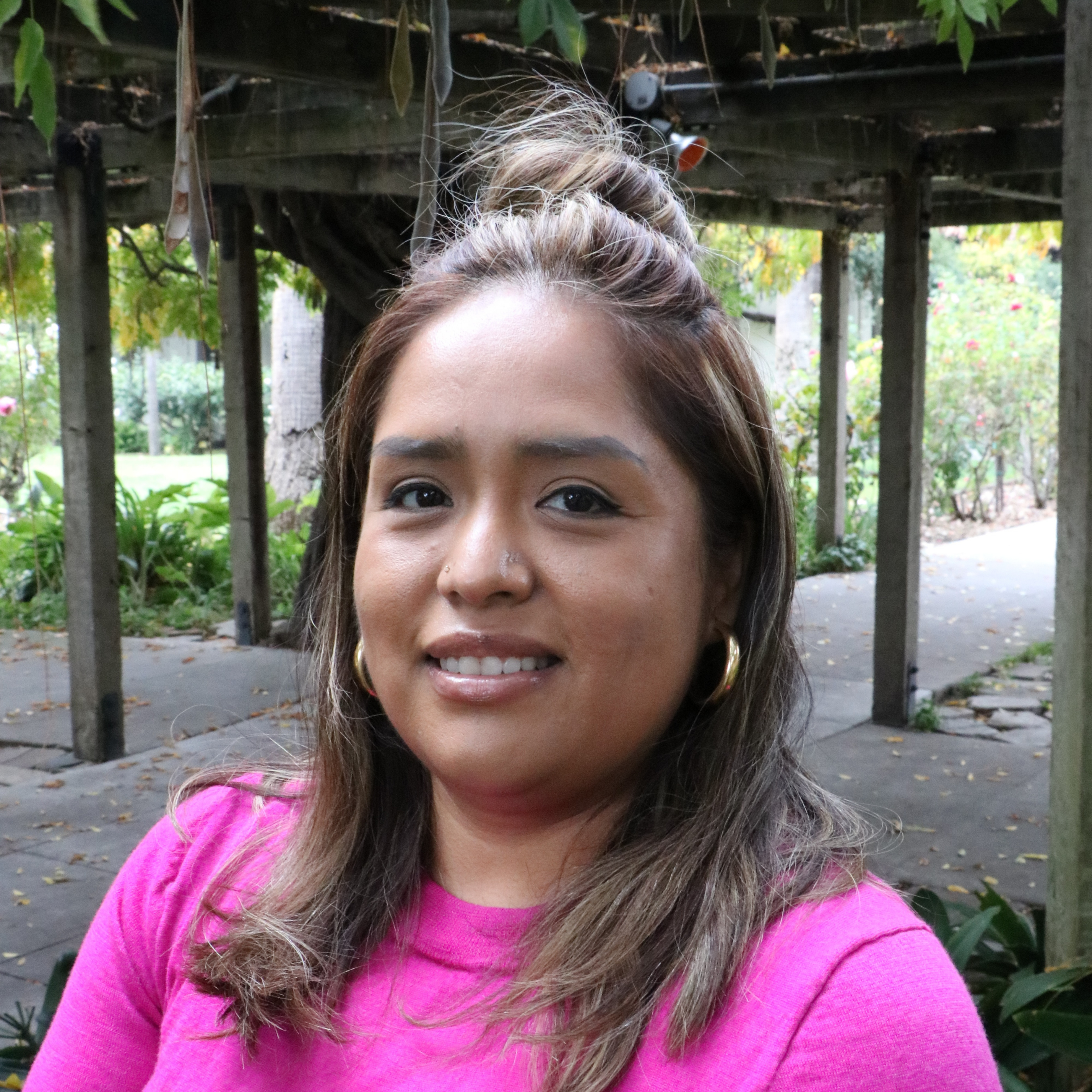 Woman in pink top smiling outdoors under a pergola at a park.