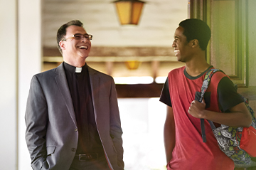 A person in a suit talking to another person holding a guitar in a hallway.