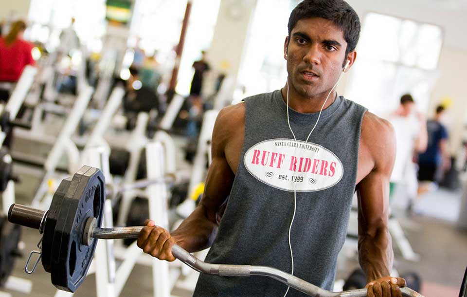Man lifting a barbell in a gym wearing a sleeveless shirt reading 