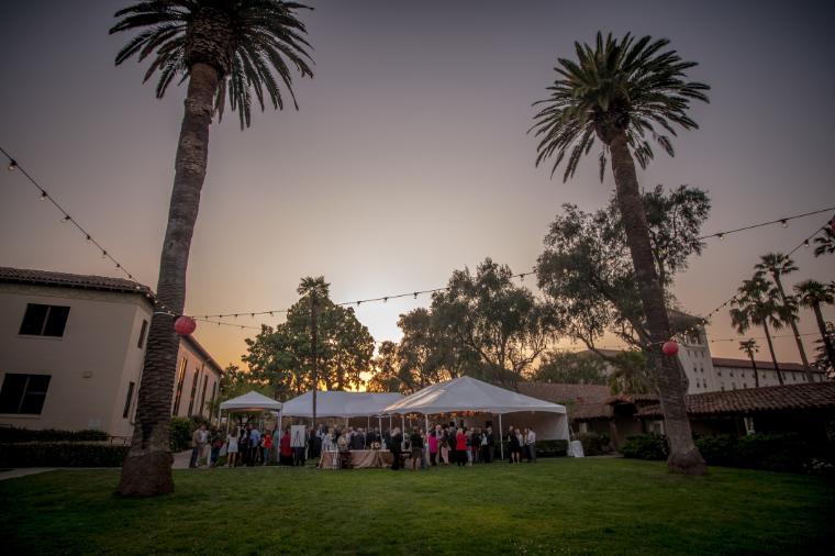 Outdoor gathering at sunset under tents, with palm trees and buildings in the background.