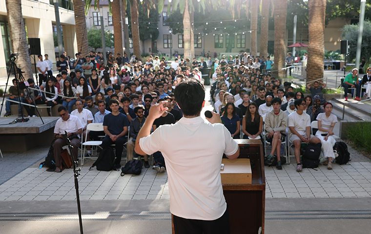 A student at a podium addresses a crowd of over 100 conference attendees in the outdoor SCDI courtyard.