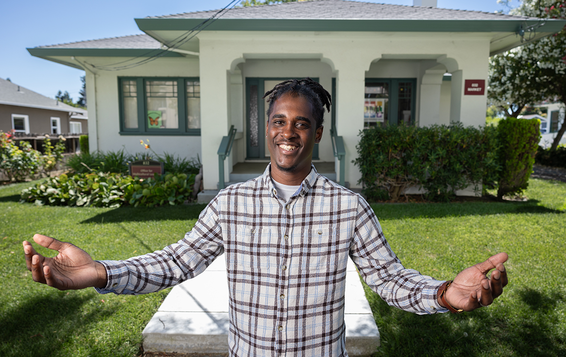 A Black man stands outside a green house, arms outstretched in a sign of welcome.