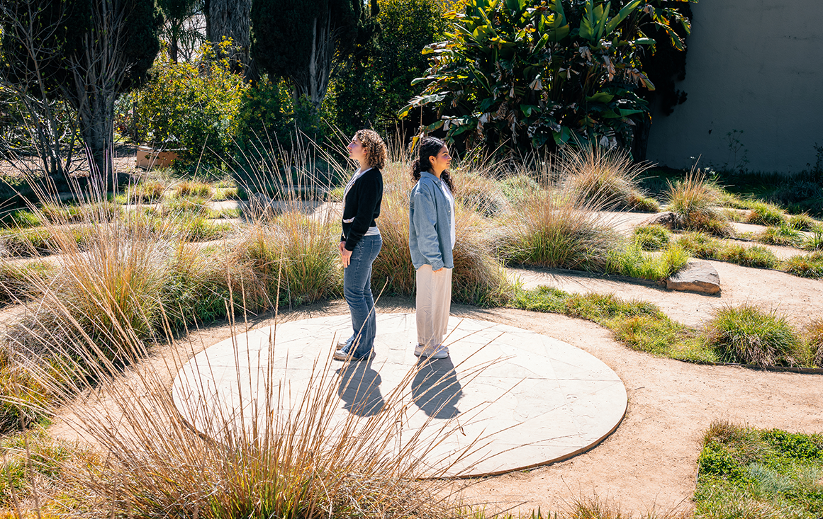 Two students stand in profile, back-to-back, in the center of an outdoor labyrinth.