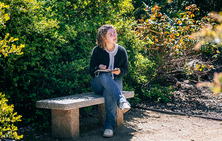 A young white woman sits on a stone bench in a garden, looking contemplative while holding a journal.