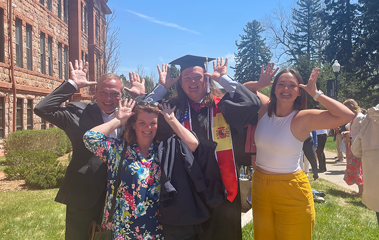 A family poses around a graduating student, holding their hands out next to their heads, thumbs pointing to their temples, fingers outstretched.
