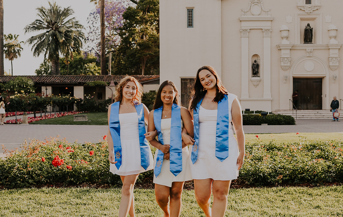 Three young women in white dresses and blue stoles walk arm-in-arm towards the camera in front of the Mission Church.