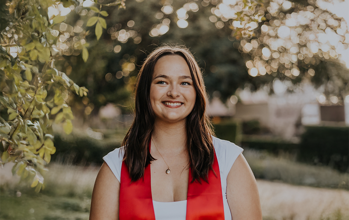 A young woman in a white shirt and red stole poses outside in the late evening.