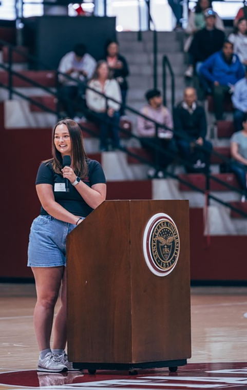 A young woman stands at a podium in a gymnasium holding a microphone and speaking to a crowd of people in the bleachers.