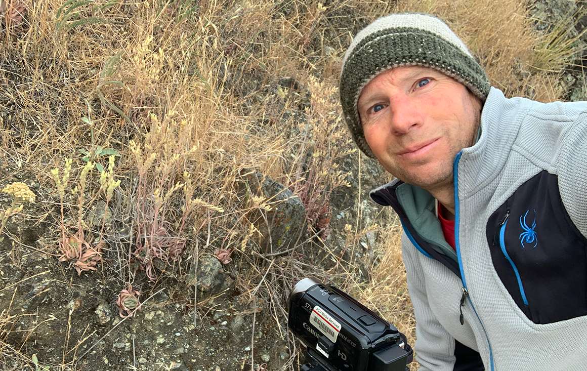 An older man in a beanie and sweater poses in a grassy field by a video camcorder which faces a succulent on a rocky outcrop.