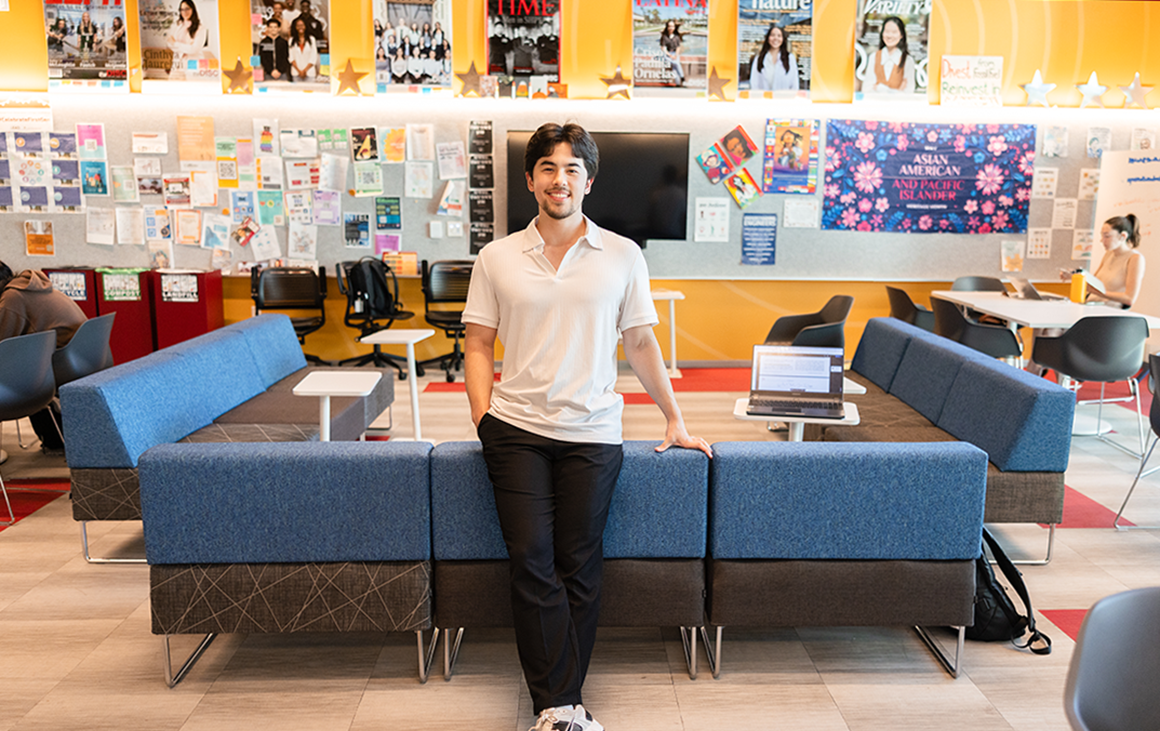 A young man leans on the back of a long couch in a brightly lit and postered lounge area.