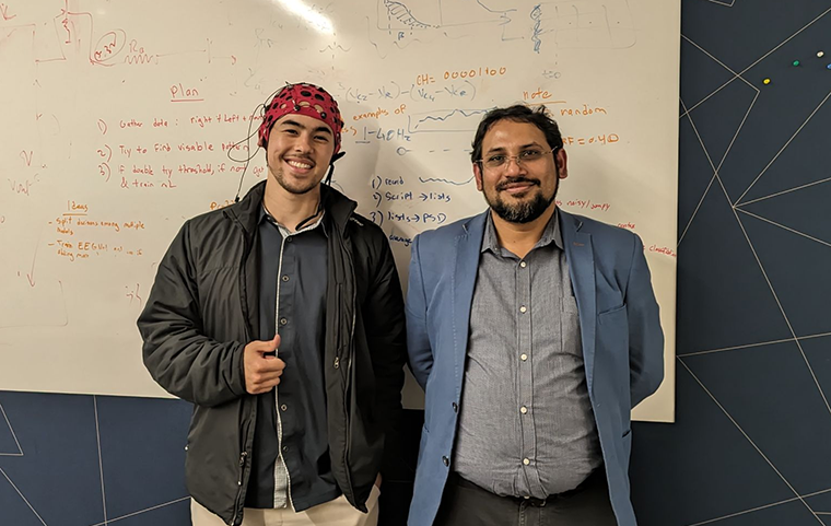 A young student wearing an EEG brain monitoring head cap stands next to any older man in front of a whiteboard.