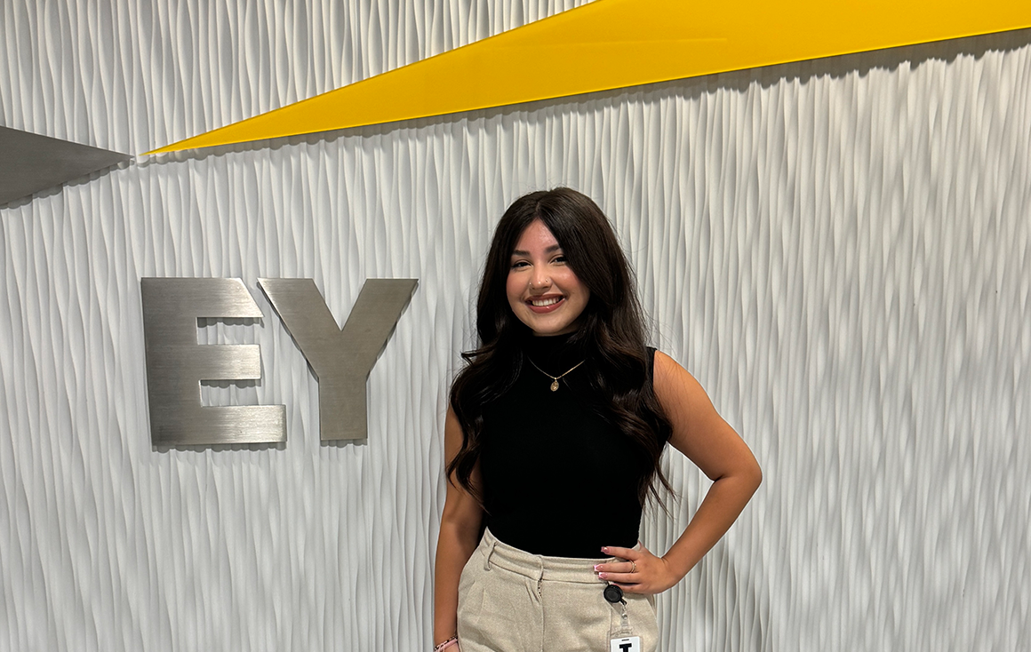 A young Latina woman stands by a corporate office sign for EY.