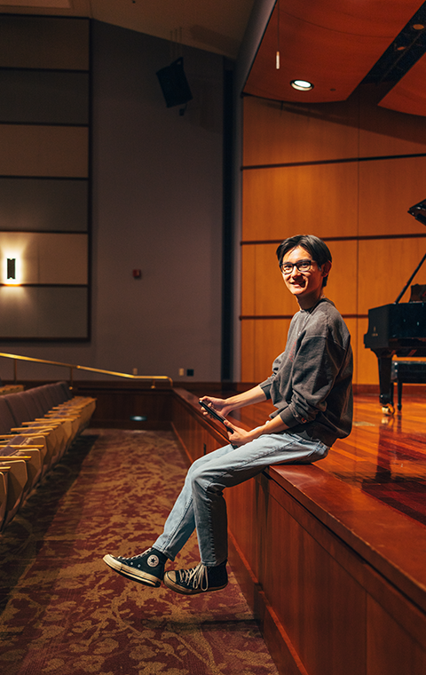 A young man sits on the edge of an empty recital hall's stage looking to the camera, holding an iPad in his hand, with a piano in the background.