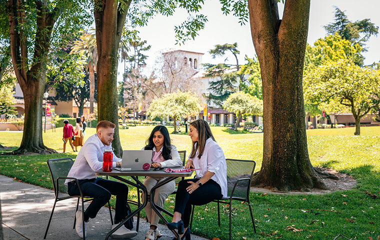 Three students outdoors at a table looking at a laptop image link to story