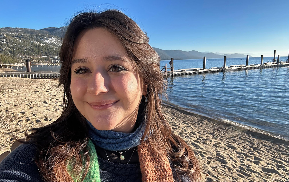 Nadine Koochou standing in a sweater on the beach in Tahoe with water and mountains in the background