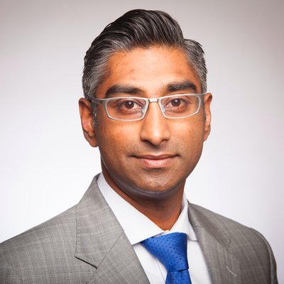 A person wearing glasses, a gray suit, and a blue tie.