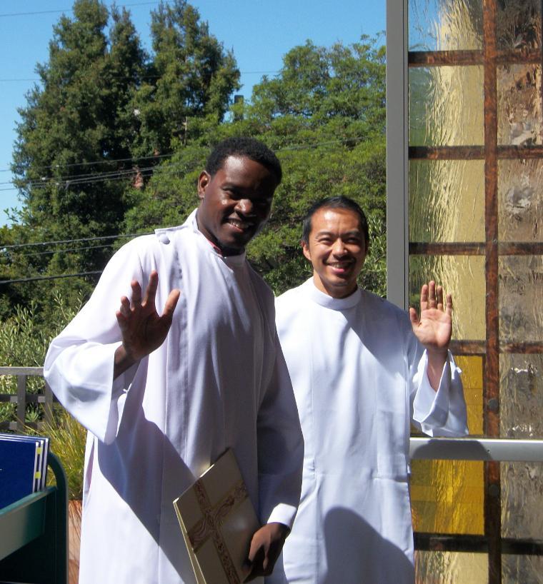 Two people in white robes smiling and waving outdoors.