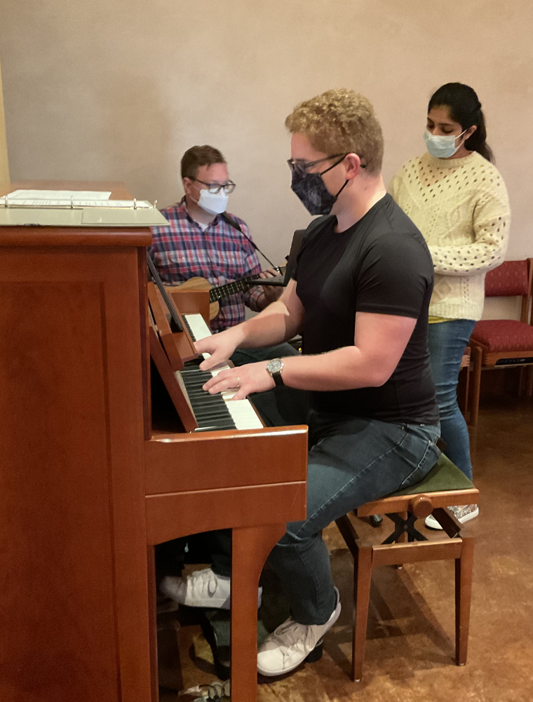 Three people, one playing piano and two standing, all wearing masks.
