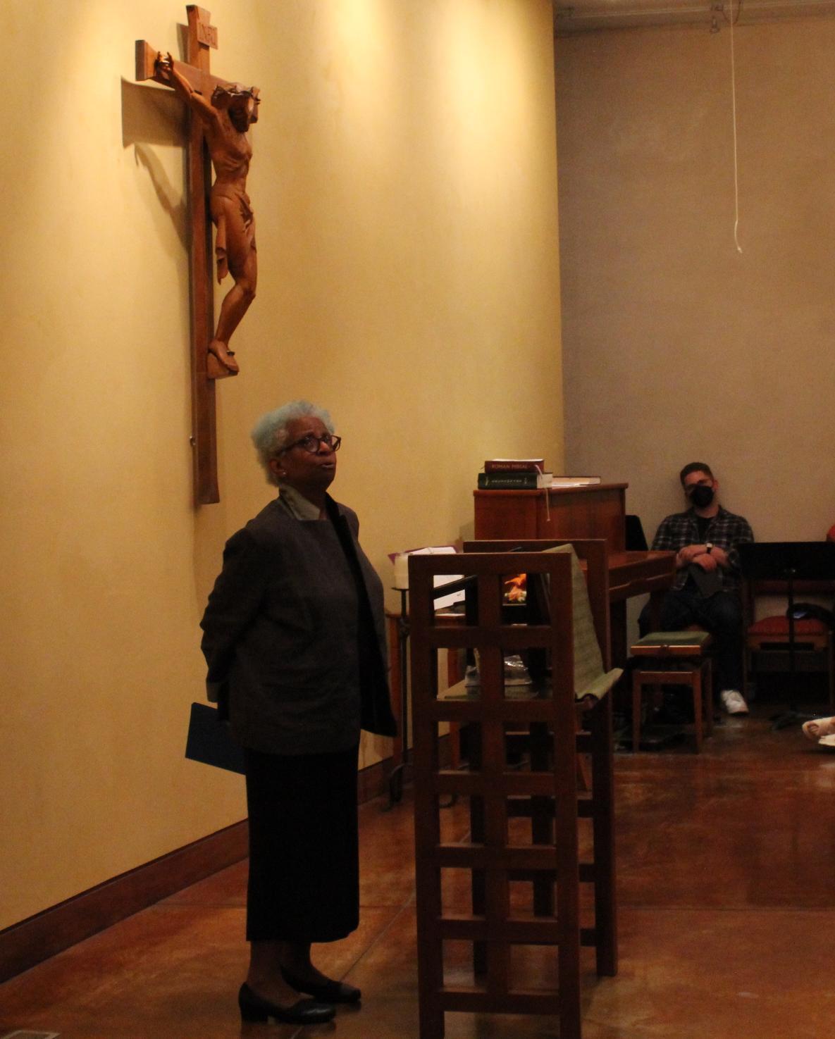 A person speaking in a room before an audience with a crucifix on the wall.