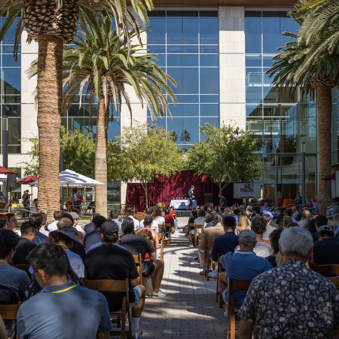 Outdoor event with people seated, a stage, and a large building backdrop.