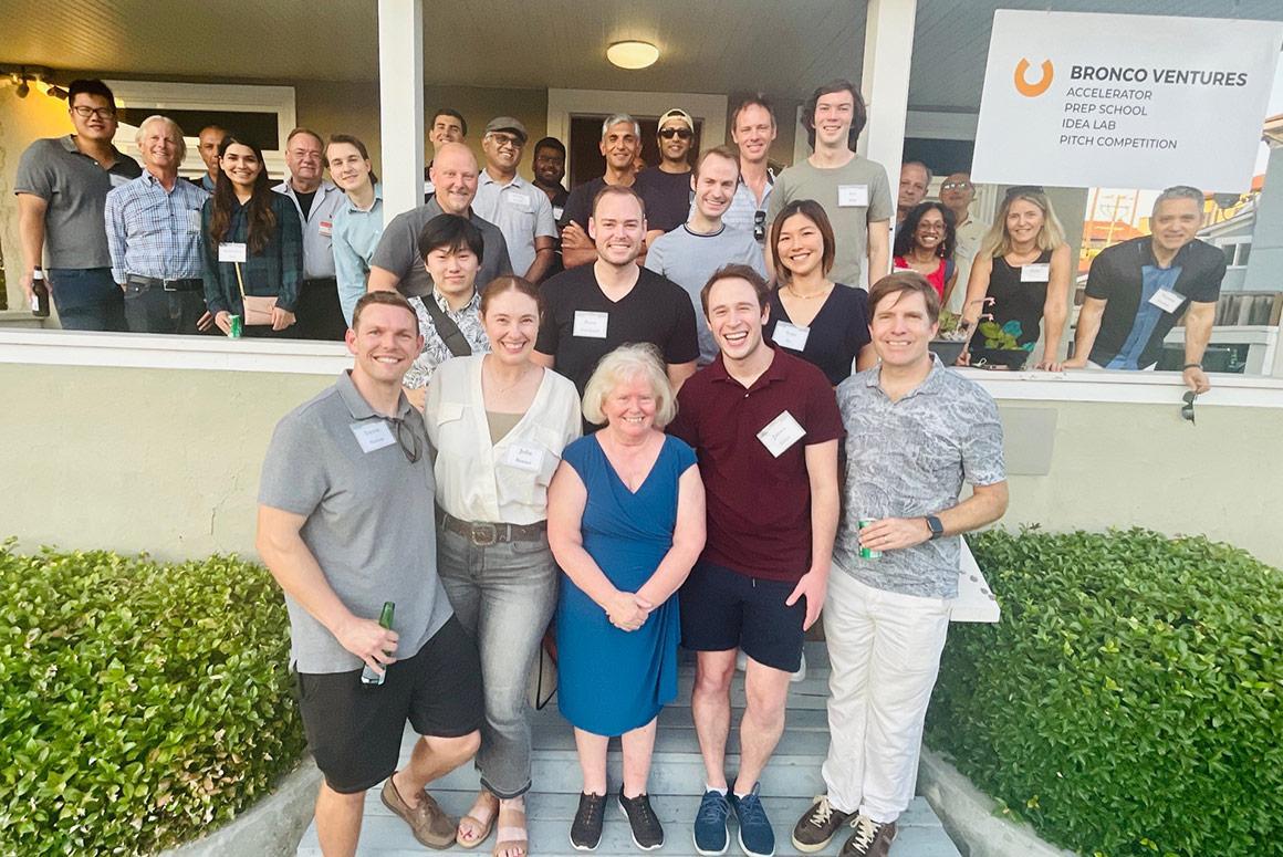 Group of people outdoors, under a porch, with a sign reading 