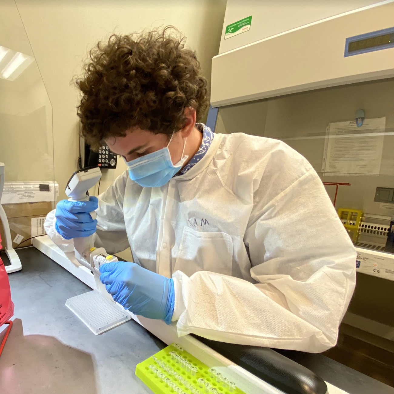 A person in a lab coat and mask working with laboratory equipment.