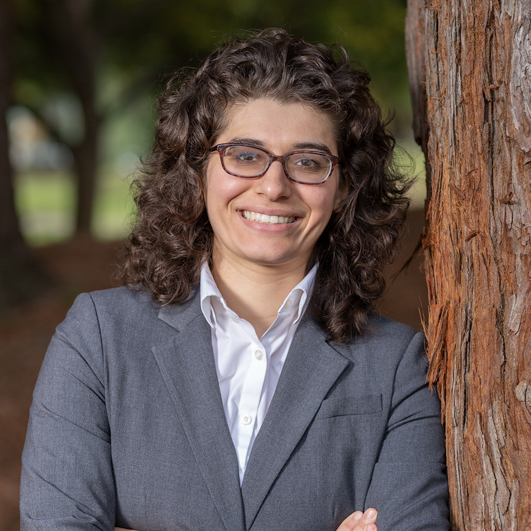 A person smiling and standing next to a tree trunk.